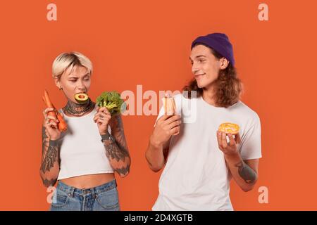 Freistil. Junge tätowierte Paar stehend isoliert auf orange Freundin hält Gemüse gesunden Lebensstil vs Freund hält Hot Dog und Donut Stockfoto