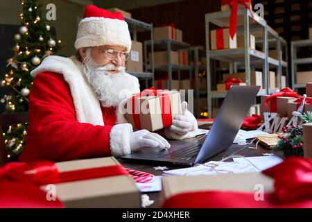 Weihnachtsmann hält Geschenk mit Laptop-Computer sitzen am Workshop-Tisch. Stockfoto