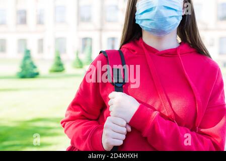 Normales Leben mit Gesichtsmaske Konzept. Cropped close up Foto Porträt von ernsthaften Mädchen in casual roten Pullover und Filter-Maske stehen in der Nähe der Schule zu bauen Stockfoto