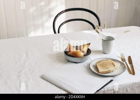 Frühstück mit Birnen Frucht Zusammensetzung. Brottoast, gefaltete Zeitungen und trockenes Gras in Becher auf weißem Leinentischtuch. Minimalistisch Skandinavisch Stockfoto