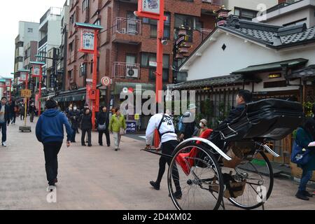 Tokio, Japan-2/28/16: Ein Arbeiter, der eine Rikscha zieht (einen Kunden trägt), während er auf eine Menge Fußgänger zuhustelt, die Asakusa hinuntergehen. Stockfoto