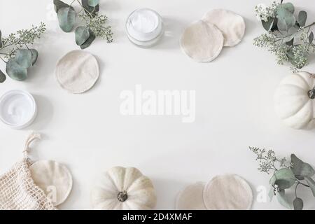 Dekorativer Kosmetikrahmen. Wiederverwendbare runde Pads aus Bio-Baumwolle zum Entfernen von Make-up. Eukalyptuszweige, Kürbisse.und Feuchtigkeitscreme auf weißem Tisch Stockfoto