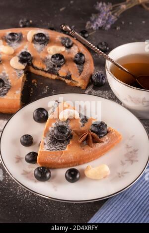 Eine in Scheiben geschnittene Torte gefüllt mit Marmelade mit Beeren und Cashewkernen. Ein Stück Kuchen mit blauen Beeren auf einer Untertasse und eine Tasse Tee mit einem silbernen Löffel auf einem dunklen ta Stockfoto