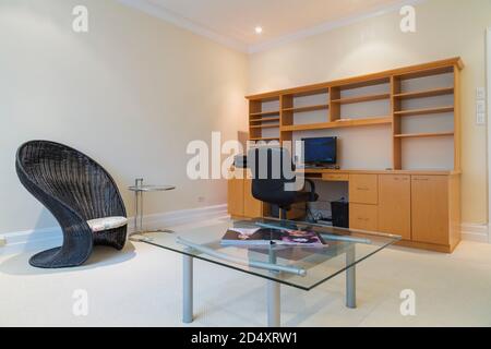 Drehsessel aus schwarzem Leder und Schreibtisch aus Holz mit Computer, Glasplatte und silbernem Metalltisch, schwarzer Korbstuhl im Home Office. Stockfoto