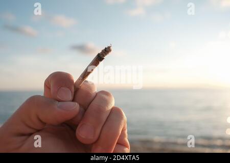 Die menschliche Hand hält eine Tabakzigarette auf dem Meer bei Sonnenuntergang Hintergrund, Raucherabhängigkeit Problem Stockfoto