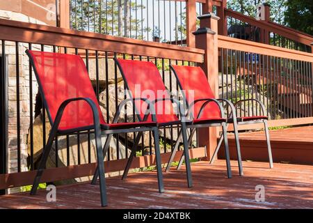 Rote Leinwandsessel auf maronfarbener Holzterrasse mit schwarzem Schmiedeeisen und Holzgeländer im Sommer. Stockfoto