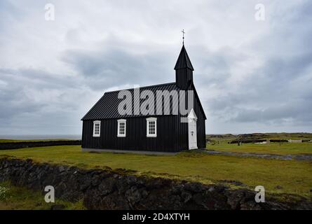 Búðakirkja, EINE schwarze Holzkirche in Budir, Snaefellsnes Peninsular, Island. Eine Steinmauer umgibt das kleine Gebäude und grüne Gras Umgebung. Stockfoto