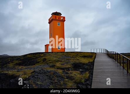 Ein Holzweg mit Geländern von der felsigen Küste bis zum orangefarbenen Svortuloft Leuchtturm auf der Snaefellsnes Halbinsel in Westisland. Stockfoto