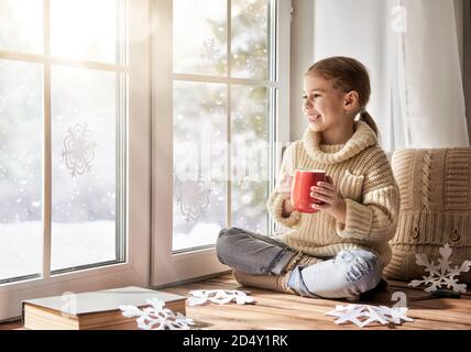 Nettes kleines Mädchen, das am Fenster sitzt, warmes Getränk trinkt und den Winterwald betrachtet. Kind macht Papier Schneeflocken für Dekoration Fenster. Stockfoto