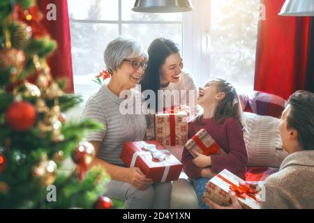Frohe Weihnachten und schöne Feiertage! Oma, Mama, Papa und Kind tauschen Geschenke aus. Eltern und Tochter haben Spaß in der Nähe von Baum drinnen. Liebevoller Familienwitz Stockfoto