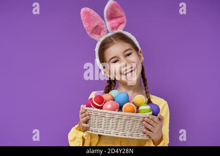 Nettes kleines Kind trägt Hase Ohren am Ostertag. Mädchen mit bemalten Eiern auf hellviolettem Hintergrund. Violette und gelbe Farben. Stockfoto