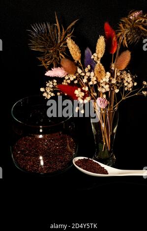Kunstfoto von Leinsamen in einem Glas und ein Löffel mit einem Bouquet von getrockneten Blumen auf einem schwarzen Hintergrund Stockfoto