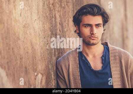 Porträt eines schönen und charmanten jungen Mannes. Er lehnt sich an eine Außenwand. Der Junge trägt einen braunen Pullover und ein blaues Hemd. Stockfoto