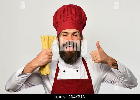 Der Küchenchef hält einen Haufen Spaghetti in der Hand. Catering und italienisches Speisekonzept. Mann mit Bart isoliert auf weißem Hintergrund. Mit gesundem Gesicht in Burgunderuniform kochen, hält trockene Pasta, die Daumen nach oben zeigen Stockfoto