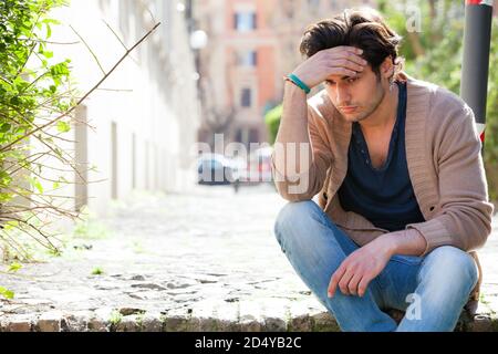 Angst. Nachdenklicher junger Mann ängstlich, im Freien. Ein junger, gutaussehender Junge sitzt auf der Straße mit einer Hand auf der Stirn. Angst, Gedanken und Stockfoto