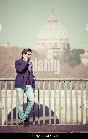 Schöner und attraktiver Kerl, modisches Kleid, Telefongespräch, während auf einer alten Brücke in Rom, Italien angehalten. Stockfoto