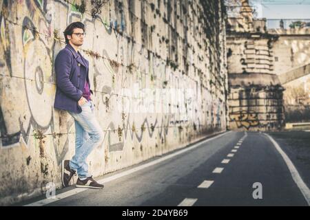 Schöner und charmanter junger Mann im Freien lehnt sich an eine Wand in der Stadt. In modischer Kleidung und mit Brille. Mantel und Jeans. Stockfoto