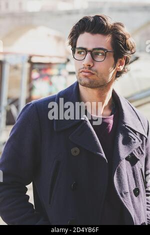 Porträt eines jungen und charmanten Mann im Freien. Der hübsche Mann trägt einen Mantel und eine Brille. Trendiger Haarschnitt. Stockfoto