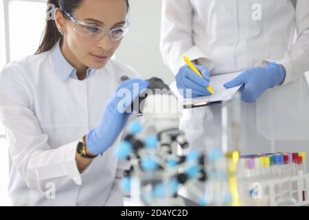 Junge Frau in weißer Uniform trägt schützende chemische Schutzbrille und Gummihandschuhe blicken ins Mikroskop Stockfoto