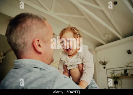 Portrait von kleinen Mädchen mit weit geöffneten Mund genießen auf Vater hebt sie in der Luft, während sie zu Hause spielt Stockfoto