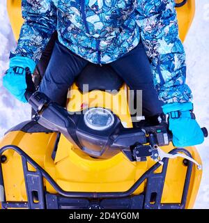 Beschnittene Nahaufnahme der Person in blauem Winteranzug und Fäustlingen, sitzend auf gelbem ATV-Fahrrad mit Helm im Winter, Draufsicht. Konzept der Winterunterhaltung, Aktivitäten und Quad-Bike. Stockfoto