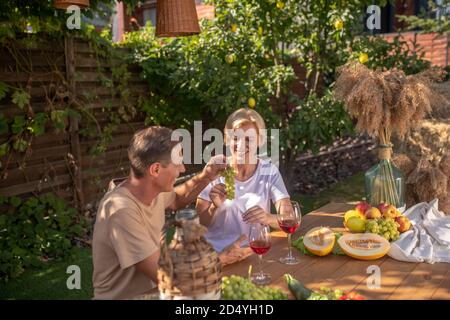 Lächelndes Paar mittleren Alters, das draußen am Tisch sitzt und Weintrauben genießt Stockfoto