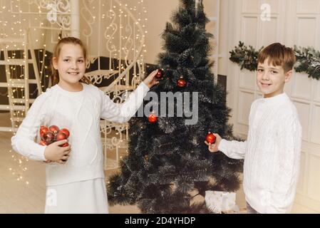 Kleine Kinder schmücken den Weihnachtsbaum in einem hellen Innenraum. Mädchen und Jungen, Bruder und Schwester hängen Neujahrsspielzeug an den Zweigen der Fichte. Familienweihnachtskonzept und Neujahrskonzept. Stockfoto