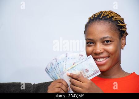 Junge afrikanische Frau hält etwas Bargeld in der Nähe ihres Gesichts und lächelt Stockfoto