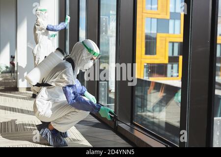 Seitenansicht Ganzkörperporträt von zwei Arbeiterinnen in Gefahrgutanzügen, die Fenster im Bürogebäude desinfizieren, Kopierraum Stockfoto