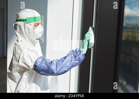 Seitenansicht Porträt einer Arbeiterin trägt Gefahrgut Anzug Desinfektion Fenster im Bürogebäude, kopieren Raum Stockfoto