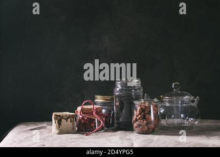 Tee- Sammlung. Schwarz trocken Tee im Glas, Rosenknospen, leere Glas Teekanne, creme Kanne, süße gezuckerte Mandeln stehen auf Tabelle mit Bettwäsche Tischdecke. Stockfoto
