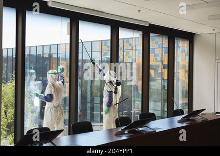 Weitwinkel-Porträt von zwei Arbeitern tragen Gefahrgut Anzüge Desinfektion Bürofenster im Konferenzraum, Kopierraum Stockfoto