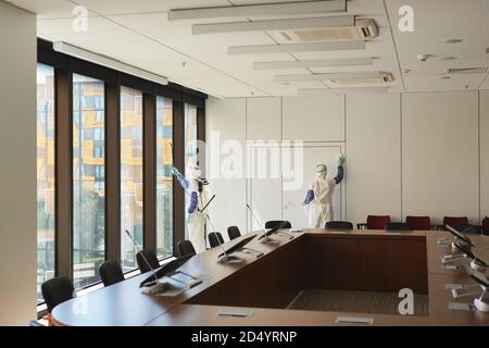 Weitwinkel-Ansicht von zwei Sanitäranlagen, die mit Gefahrgutanzügen ausgestattet sind, die den Konferenzraum im Büro desinfizieren, Kopierbereich Stockfoto