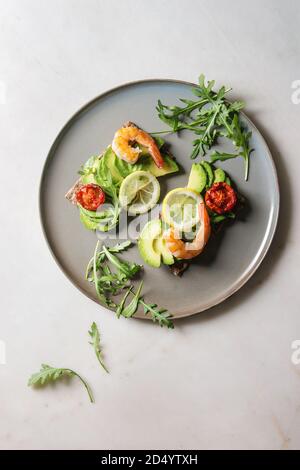 Vegetarisches Sandwich mit in Scheiben geschnittenen Avocado, sonnengetrocknete Tomaten, Garnelen, Rucola, serviert auf keramische Platte über weißem Marmor Hintergrund. Flach, Raum Stockfoto