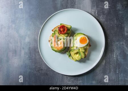 Vegetarisches Sandwich mit in Scheiben geschnittenen Avocado, sonnengetrocknete Tomaten, Ei, Garnelen, auf keramische Platte über blaue Textur Hintergrund serviert. Flach, Raum Stockfoto