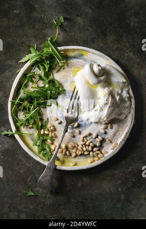 Geschnetzeltes Italienische burrata Käse, frische Rucola Salat, Pinienkerne und Olivenöl in weißer Keramik Platte über Dark Metal Hintergrund. Flach, Raum Stockfoto