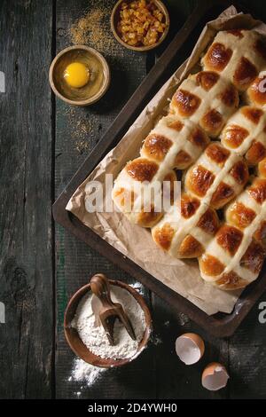 Hausgemachte Ostern traditionelle Hot Cross Buns auf ein Backblech mit Backpapier auslegen und Zutaten oben auf dunklem Hintergrund. Ansicht von oben, Platz. Rustikale s Stockfoto