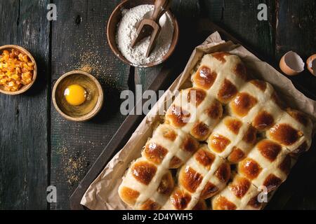 Hausgemachte Ostern traditionelle Hot Cross Buns auf ein Backblech mit Backpapier auslegen und Zutaten oben auf dunklem Hintergrund. Ansicht von oben, Platz. Rustikale s Stockfoto