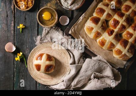 Hausgemachte Ostern traditionelle Hot Cross Buns auf Platte und ein Backblech mit Backpapier auslegen und Zutaten oben auf dunklem Hintergrund. Ansicht von oben, Raum Stockfoto