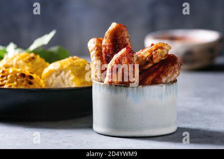 BBQ spicy Chicken Wings in Keramik Schüssel mit roter Soße serviert, gegrillter Zuckermais und Petersilie Bündel auf hellblau Tabelle. Stockfoto