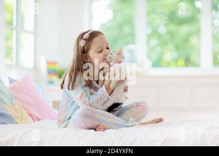 Kind spielt mit Baby Katze auf dem Bett in weissen Schlafzimmer. Kid Holding weiße Katze. Kleines Mädchen im Schlafanzug mit niedlichen Haustier zu Hause. Kinder spielen mit cat Stockfoto