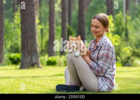 Nahaufnahme der lächelnden Frau in kariertem Hemd umarmt und umarmt mit Zärtlichkeit und Liebe Hausginger Katze, streichelt auf dem Kopf, im Freien in der Sonne Stockfoto