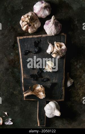 Vielzahl von frischen Bio Knoblauch Zwiebeln und Knoblauchzehen geschält und schwarze fermentierte Knoblauch auf Holz Schneidebrett über Dark Metal Hintergrund. Flach Stockfoto