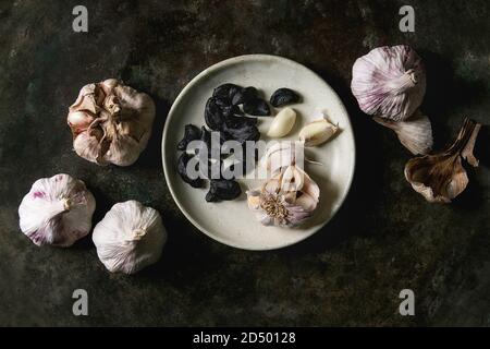 Vielzahl von frischen Bio Knoblauch Zwiebeln und Knoblauchzehen geschält und schwarze fermentierte Knoblauch auf keramische Platte über Dark Metal Hintergrund. Flach, Spa Stockfoto