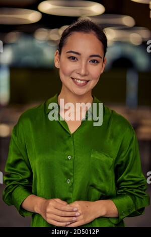 Vertikale Aufnahme einer jungen glücklichen asiatischen Geschäftsfrau in grünem Seidenhemd, die Kamera anschaut und lächelt, während sie im Büro posiert. Business People-Konzept Stockfoto