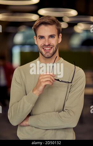 Vertikale Aufnahme eines jungen glücklichen kaukasischen Geschäftsmannes, der seine Brille hält, die Kamera anschaut und lächelt, während er im Büro posiert. Business People-Konzept Stockfoto