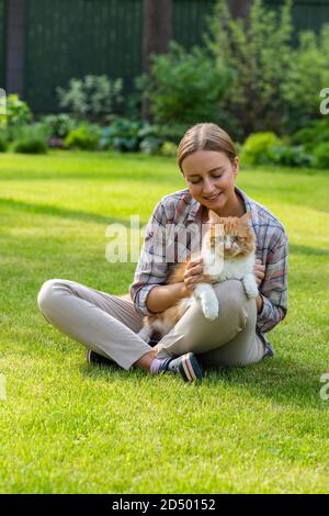 Nahaufnahme der lächelnden Frau in kariertem Hemd umarmt und umarmt mit Zärtlichkeit und Liebe Hausginger Katze, streichelt auf dem Kopf, im Freien in der Sonne Stockfoto