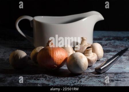 Stillleben Bild von rohen Pilzen, Zwiebel, Messer und Soße Boot über dunklen Hintergrund Stockfoto