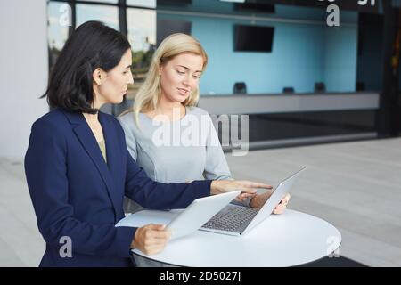 Waist-up-Porträt von zwei erfolgreichen Geschäftsfrauen diskutieren Arbeit, während am Schreibtisch im Bürogebäude stehen und mit Laptop, Kopierer Platz Stockfoto