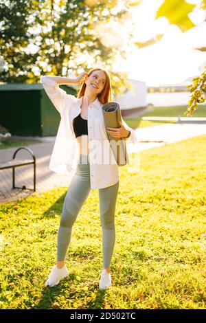 Porträt einer fröhlichen attraktiven rothaarigen jungen Frau mit Yogamatte, im Stadtpark stehend. Stockfoto